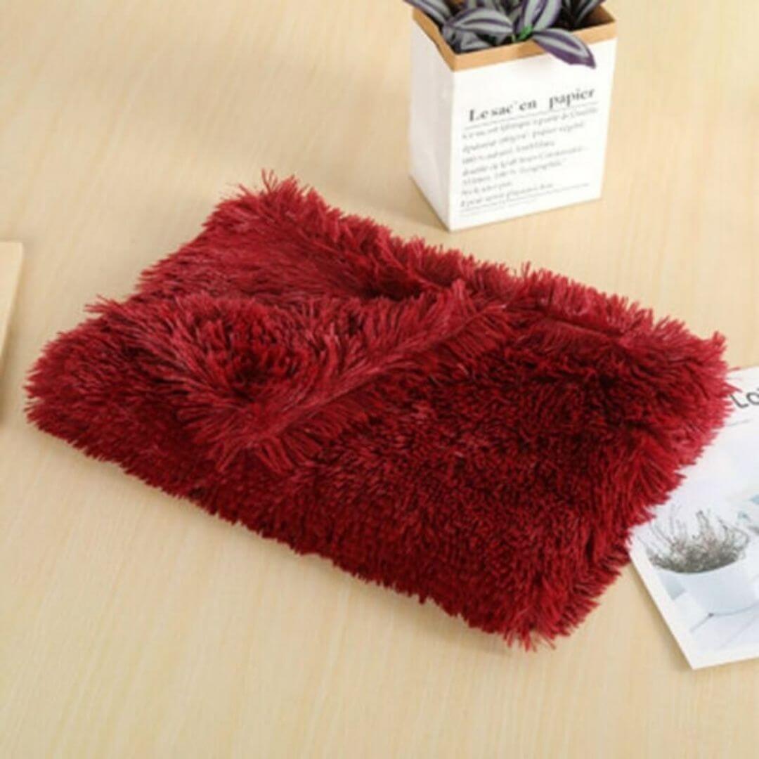 A soft, plush wine red anxiety relieving blanket folded neatly on a wooden table beside a small potted plant and some documents